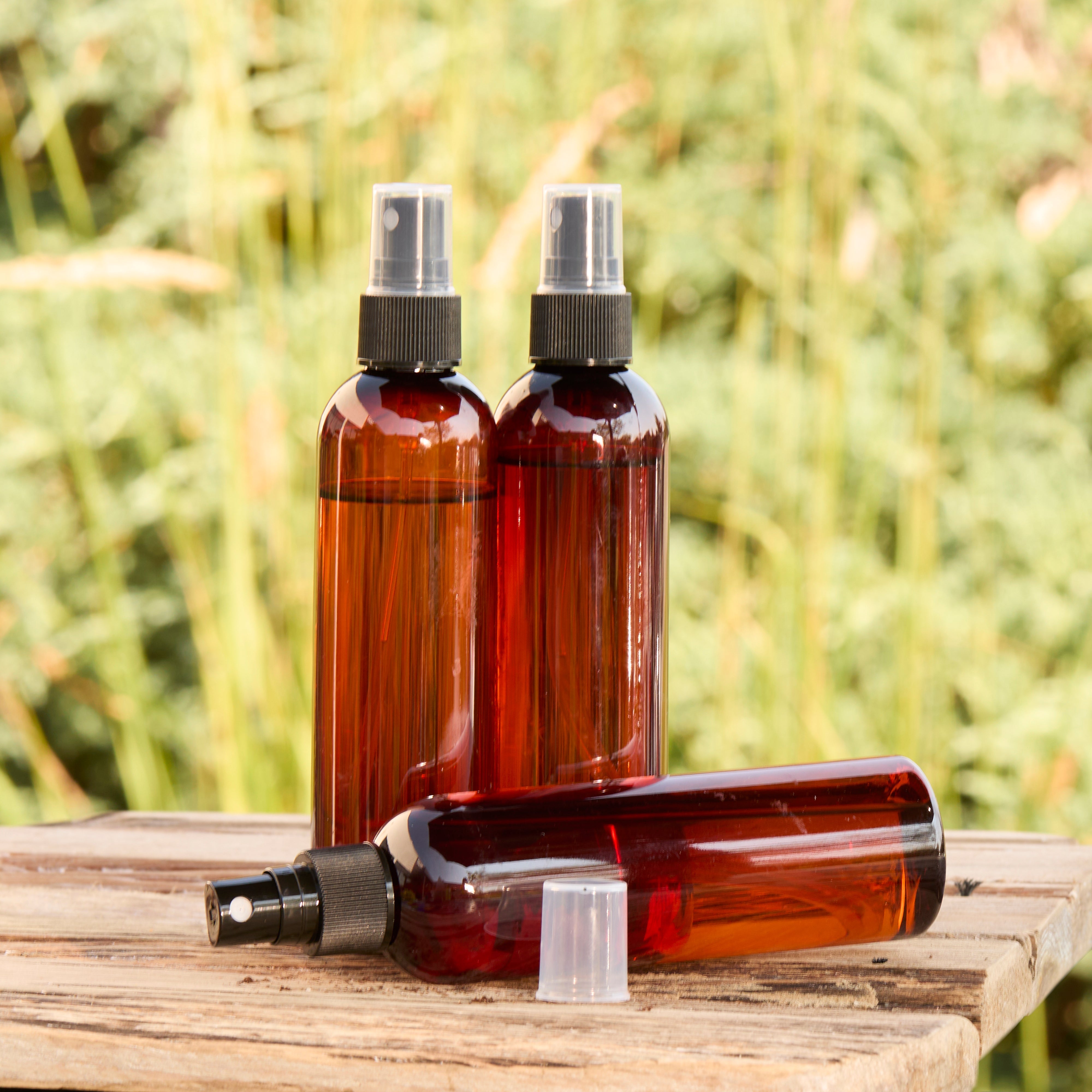 Three amber plastic spray bottles with black sprayers are displayed on a wooden surface outdoors, with green foliage blurred in the background. Two bottles stand upright and one lies on its side with the clear cap removed and placed nearby.