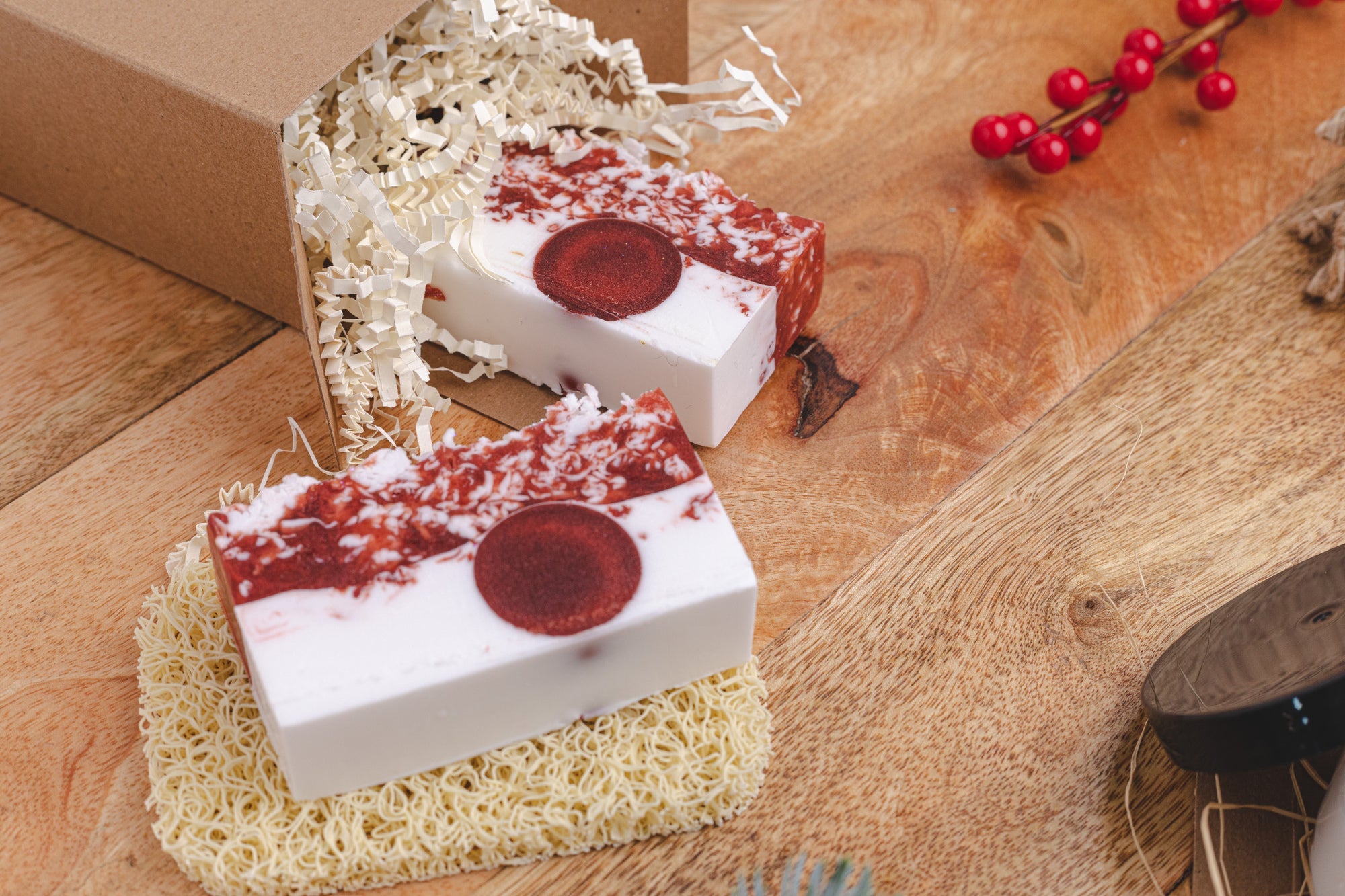 Handcrafted soap bars with red and white layers and a circular embed, styled on a wooden surface with natural packing filler, a kraft gift box, and festive red berry accents.