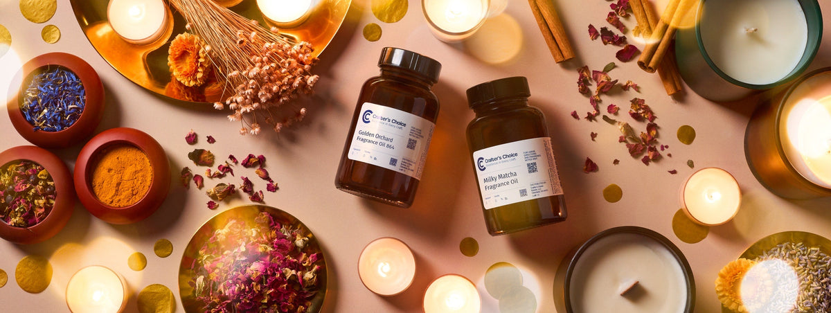 Two amber bottles of Crafter’s Choice fragrance oils, Golden Orchard and Milky Matcha, surrounded by glowing candles, dried flowers, cinnamon sticks, colorful botanicals, and gold confetti on a soft pink background.