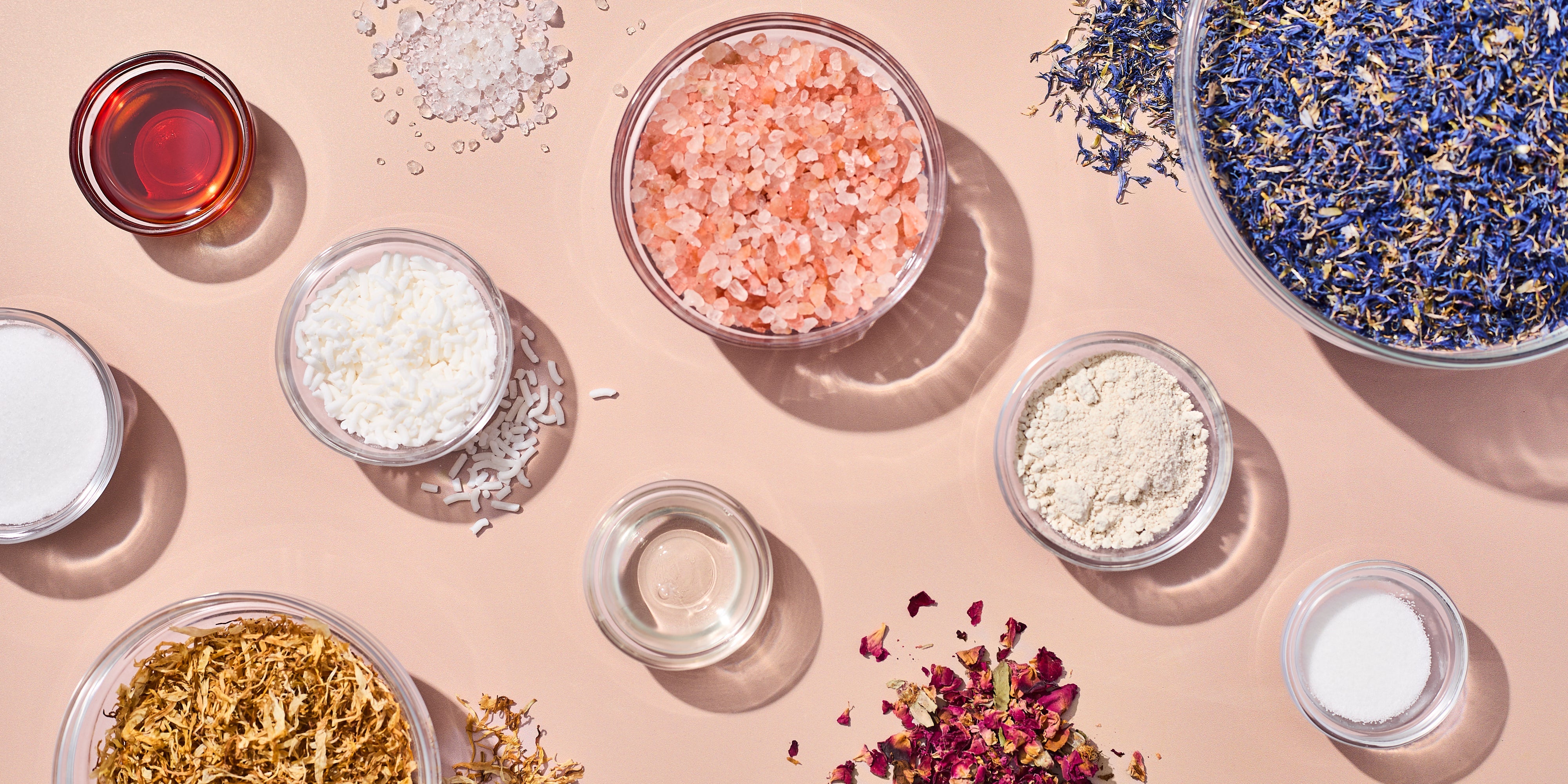 Flat lay of bath and body additives on a blush surface, including pink Himalayan salt, dried botanicals, clay powder, wax pellets, liquid additives, and fine salts arranged in clear bowls and small piles.