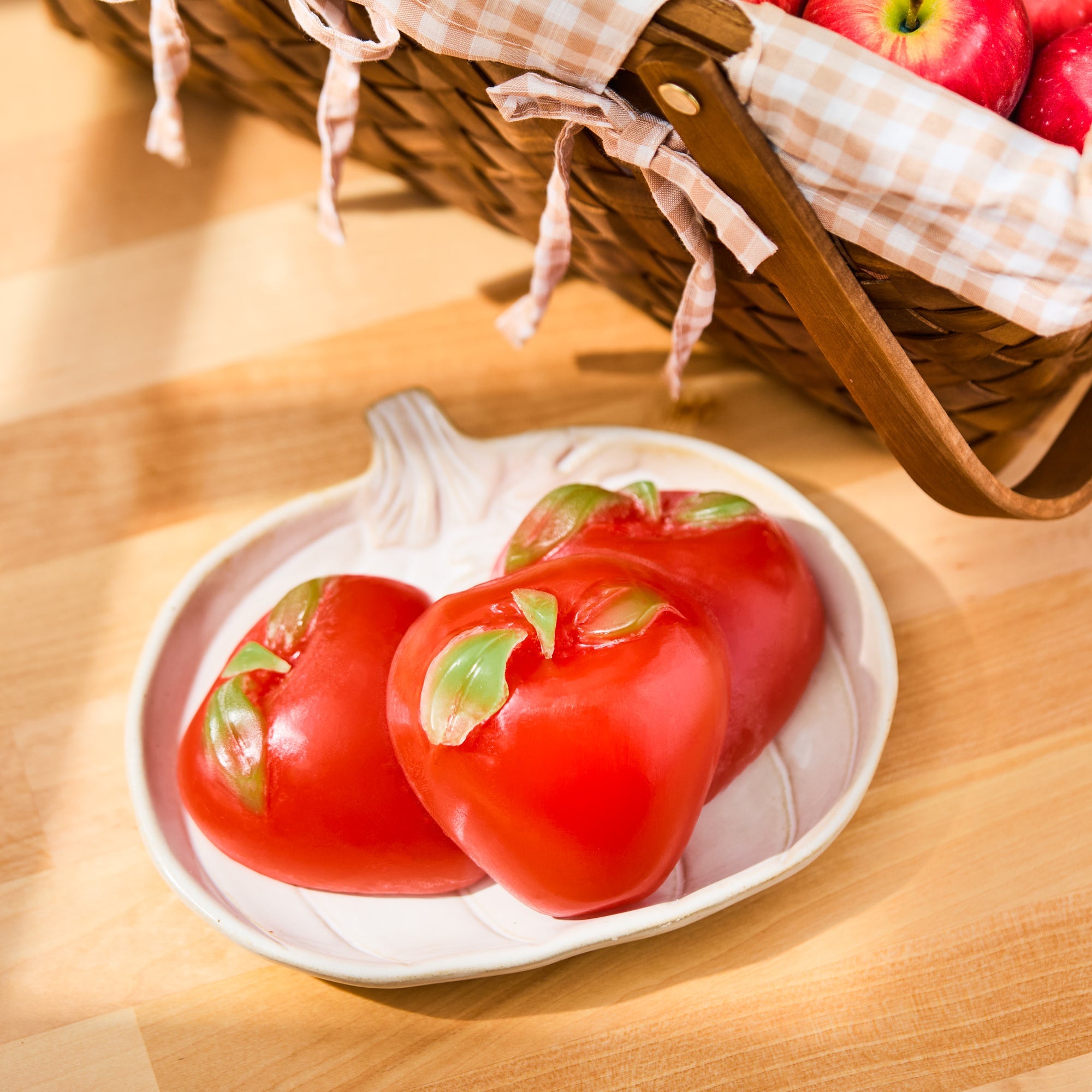 Red apple shaped soaps with green leaves displayed on white dish beside wicker basket of apples