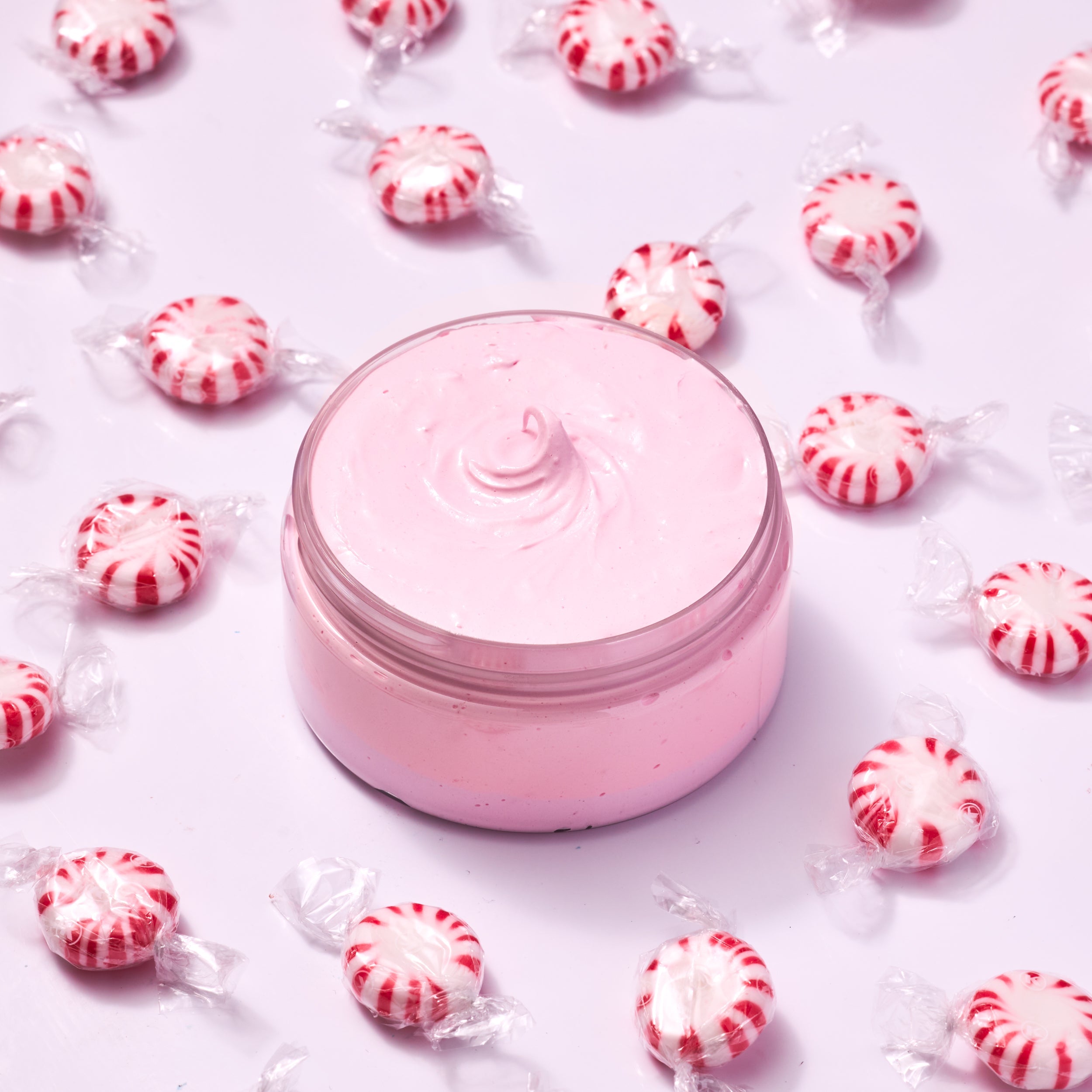 A clear round jar filled with smooth, pastel pink whipped cream or scrub sits centered on a white background, surrounded by individually wrapped red-and-white peppermint candies arranged in a scattered pattern.