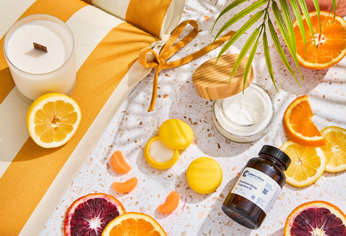 Bright citrus-themed flat lay featuring a white candle, jar of body cream, yellow lip balm containers, and a fragrance oil bottle surrounded by sliced lemons, oranges, blood oranges, and citrus segments on a terrazzo surface.