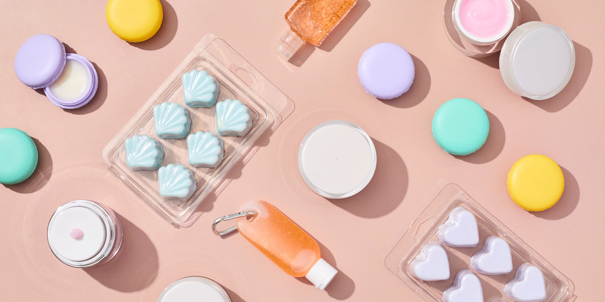 Flat lay of colorful cosmetic packaging including macaron-style containers, small jars, clamshell molds filled with shell and heart shapes, and a mini orange squeeze bottle on a soft peach background.