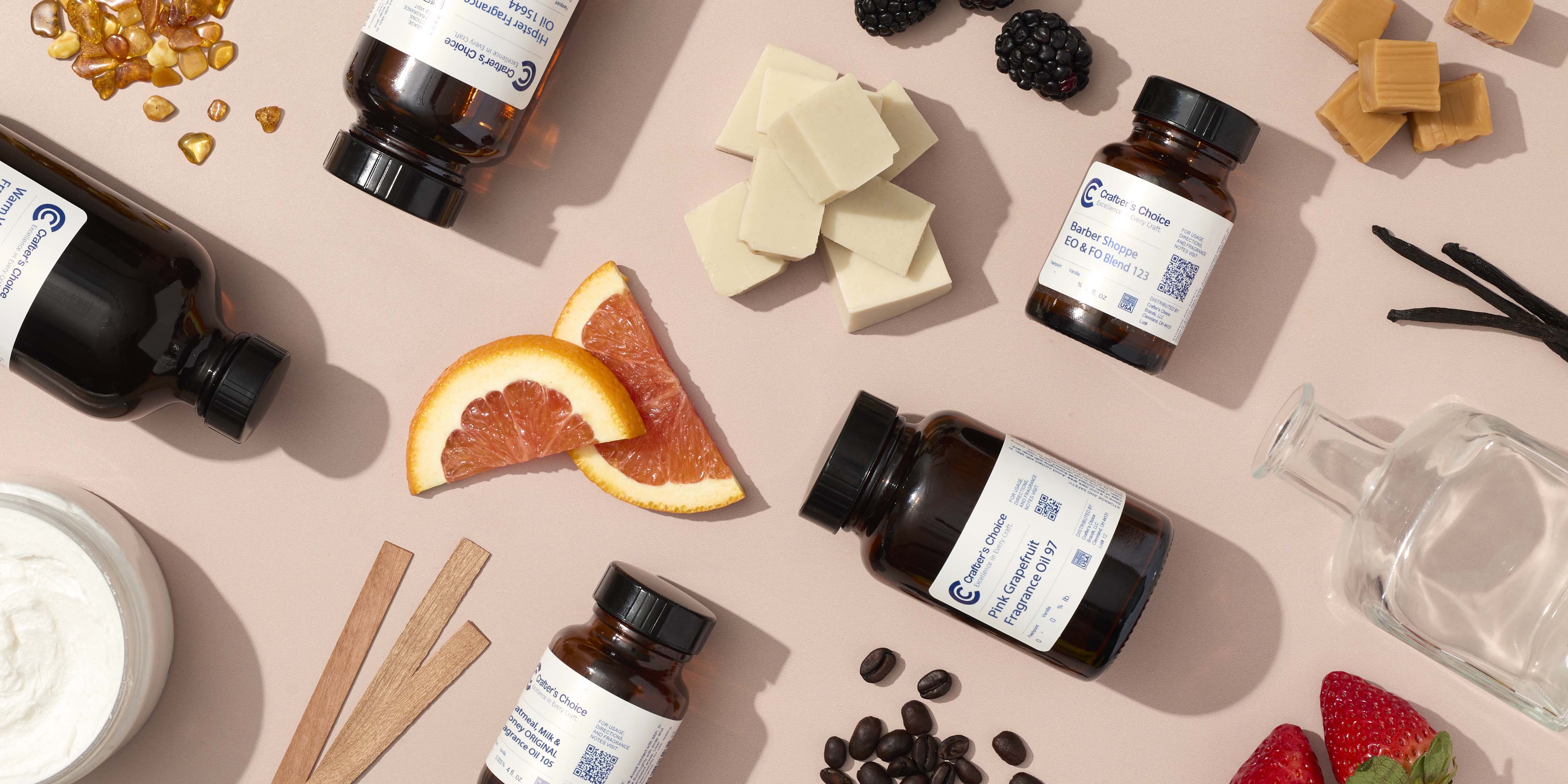 Flat lay of fragrance oil bottles on a soft pink background, styled with scent elements like citrus slices, berries, vanilla beans, caramel pieces, wax blocks, wood strips, coffee beans, and amber chips.