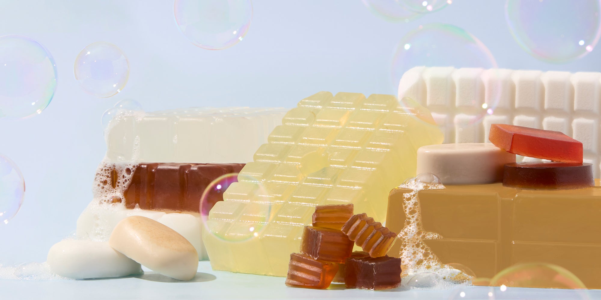 Melt and pour soap bars and embeds in assorted colors arranged with foam and floating bubbles on a light blue background.