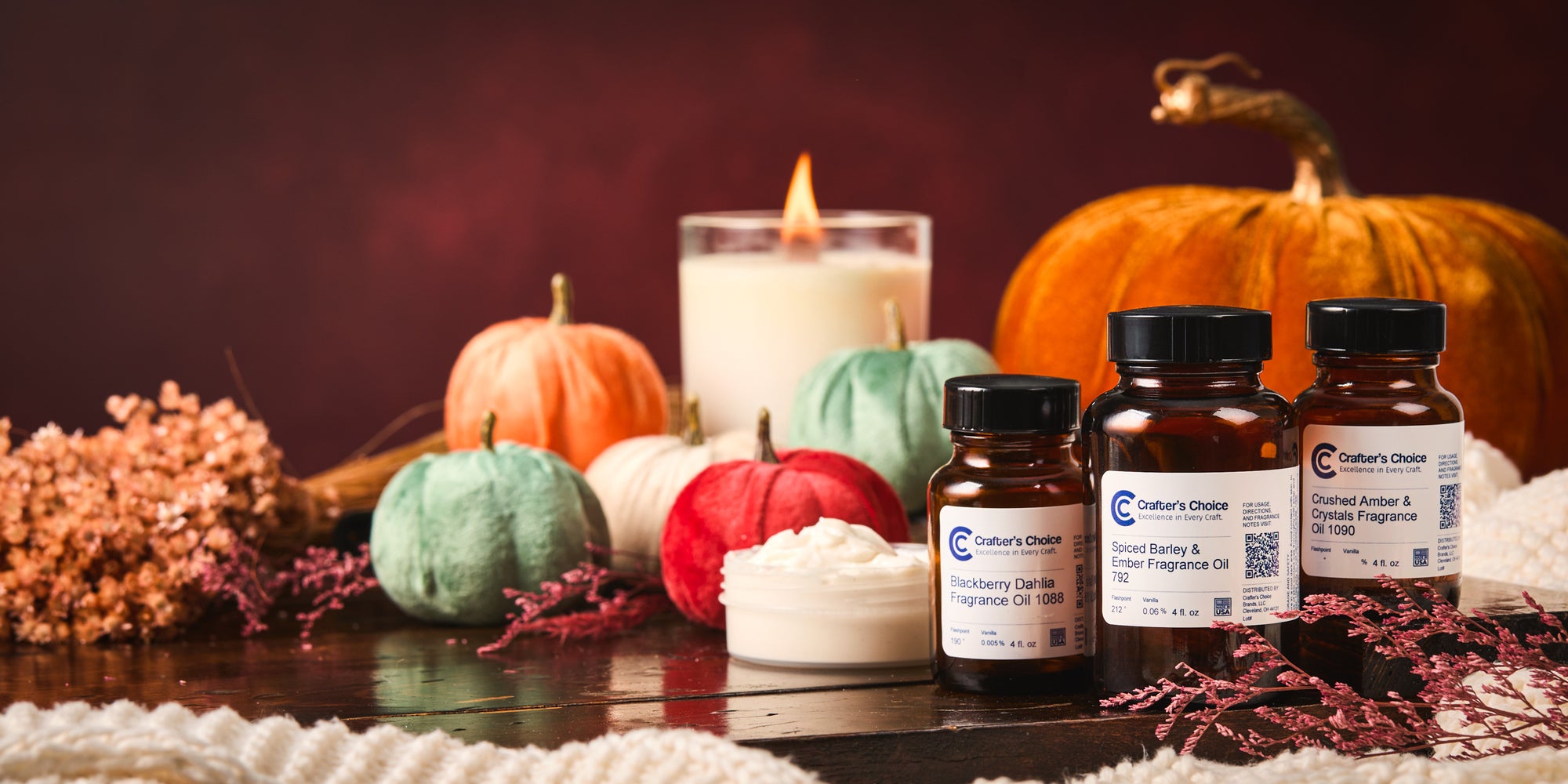 Autumn-themed display featuring four amber bottles of Crafter's Choice fragrance oils, a jar of white whipped body butter, colorful mini pumpkins, a lit candle, dried florals, and a large pumpkin on a rustic wooden surface.
