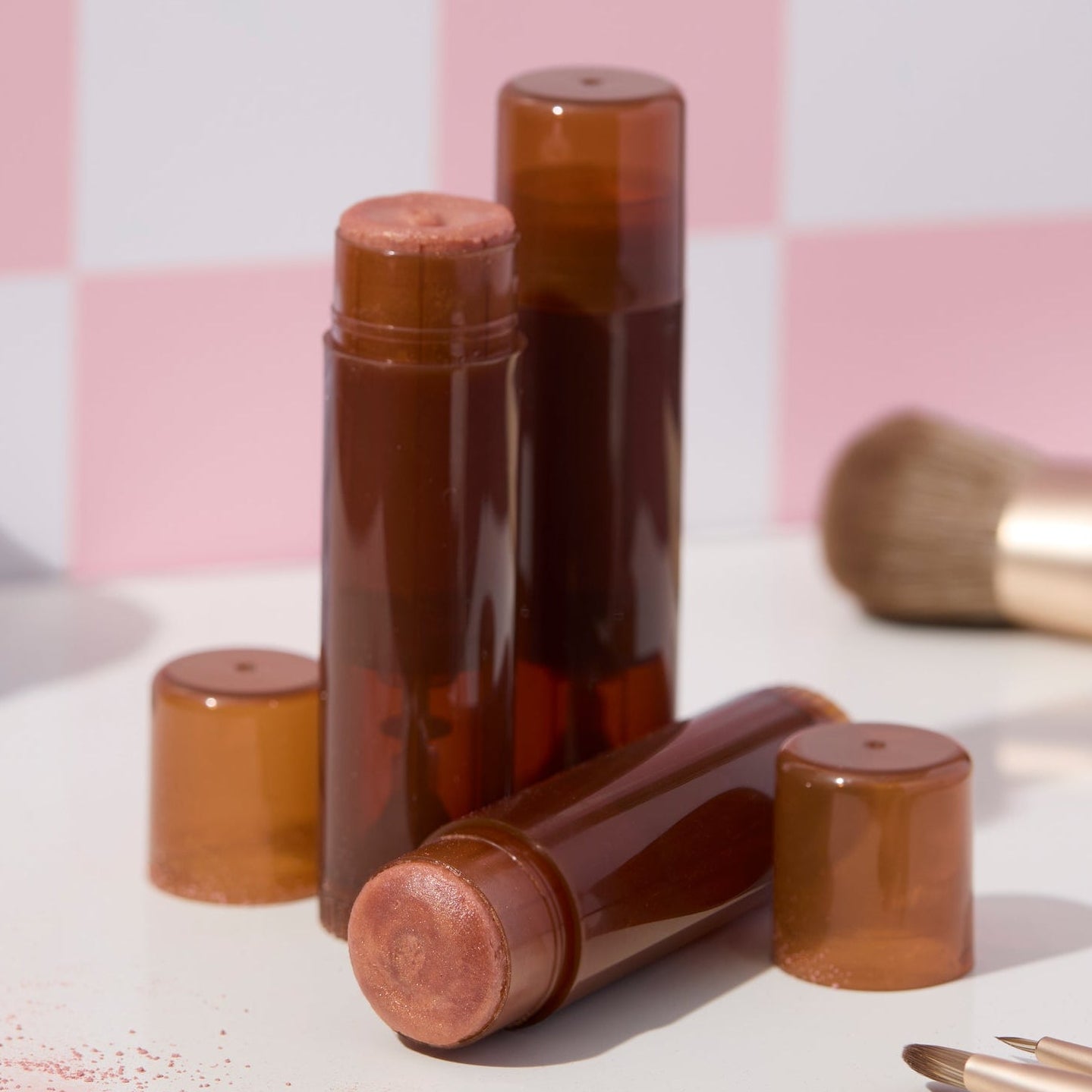 Golden shimmer makeup sticks in amber tubes displayed on a white surface, one upright and one lying down, with soft pink powder visible at the tips. Makeup brushes and loose shimmer powder sit around them against a pink checkered background.