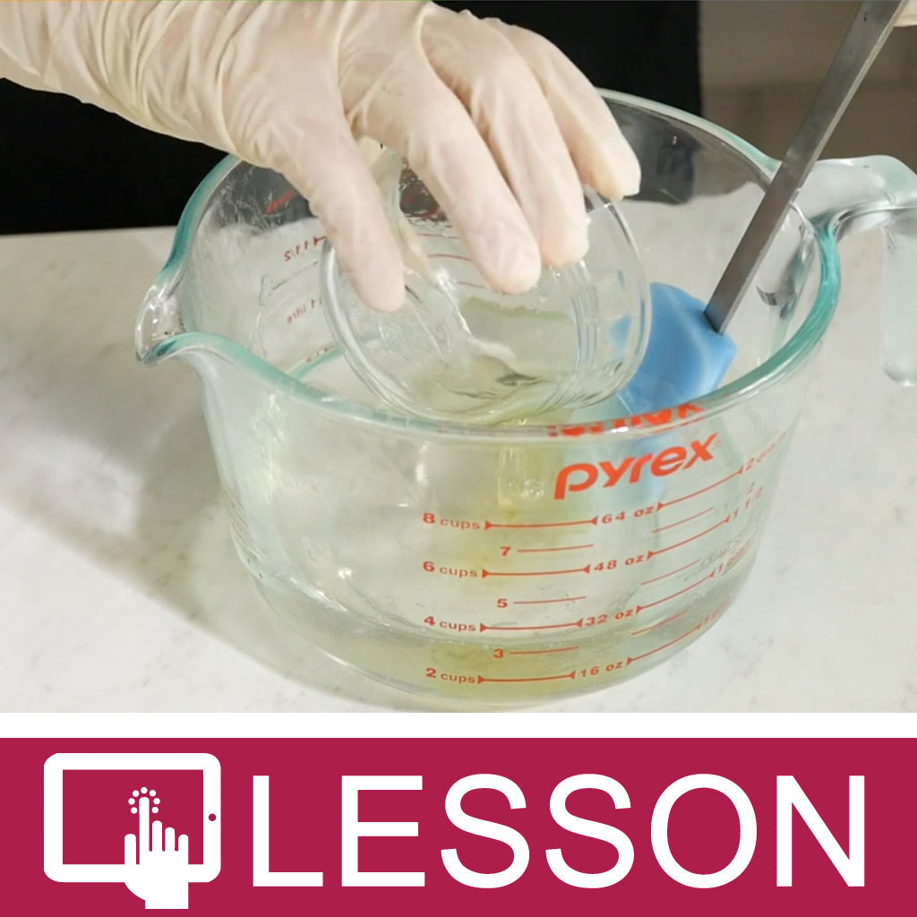 Gloved hand adding liquid to melted soap base in a glass measuring jug