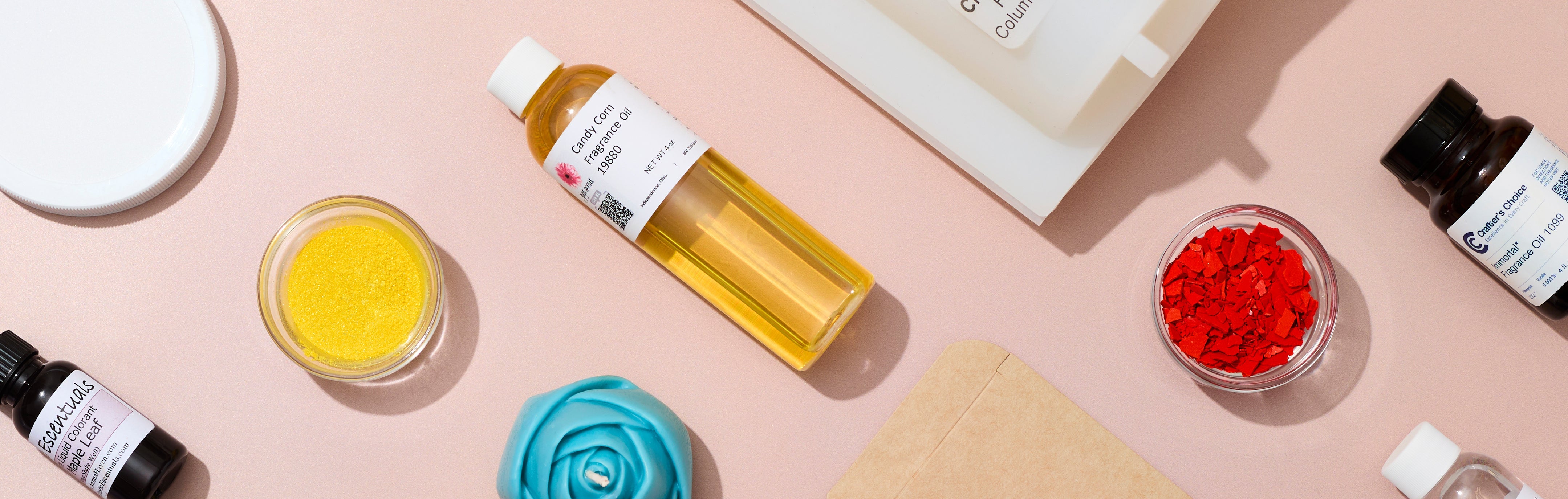 Assorted soap-making and fragrance ingredients arranged on a soft pink background, including fragrance oil bottles, yellow mica powder, red color flakes, a blue rose-shaped soap, and packaging materials in a flat-lay composition.