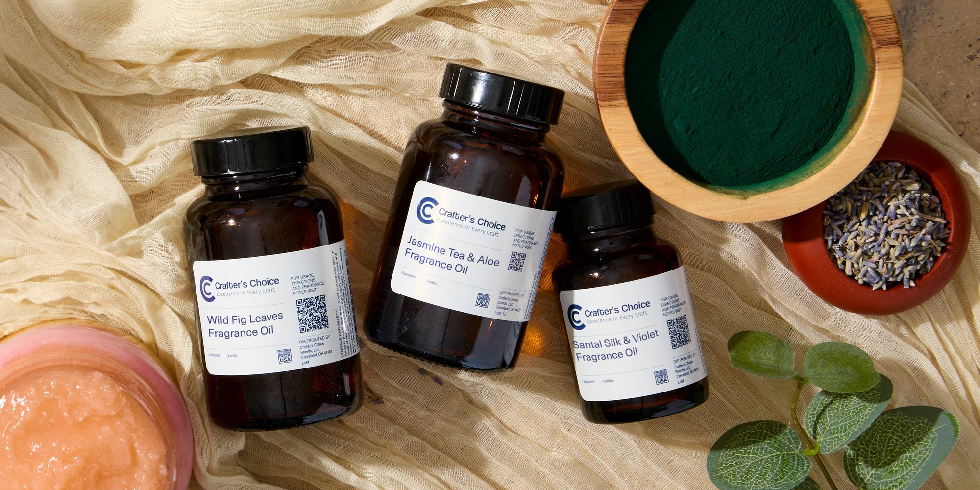 Three amber bottles of Crafter’s Choice fragrance oil—Wild Fig Leaves, Jasmine Tea & Aloe, and Santal Silk & Violet—arranged on beige fabric beside bowls of green powder and dried lavender, with leaves and a pink gel mixture nearby.